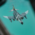 Aerial view of a military fighter jet soaring above vibrant turquoise waters.