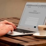 Close-up of hands typing on a laptop next to a cup of coffee on a wooden desk.